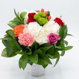 Vibrant mixed floral arrangement with pink carnations, orange and purple roses, and green chrysanthemums in a white square vase.